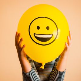 Ein Luftballon mit einem Smiley wird vor das Gesicht gehalten. 