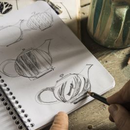 Blick auf eine Hand beim Zeichnen einer Teekanne in einem Notizbuch.