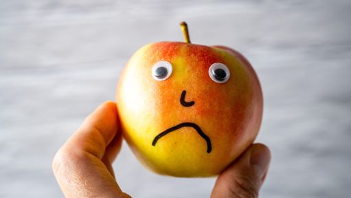 Eine Hand hält einen Apfel mit aufgeklebten Wackelaugen und aufgemalter Nase und traurigem Mund