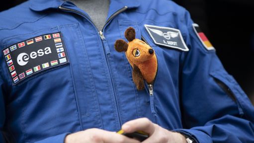 Ein blauer ESA-Fluganzug von Alexander Gerst mit einer Plüschfigur der „Maus" in der Brusttasche.