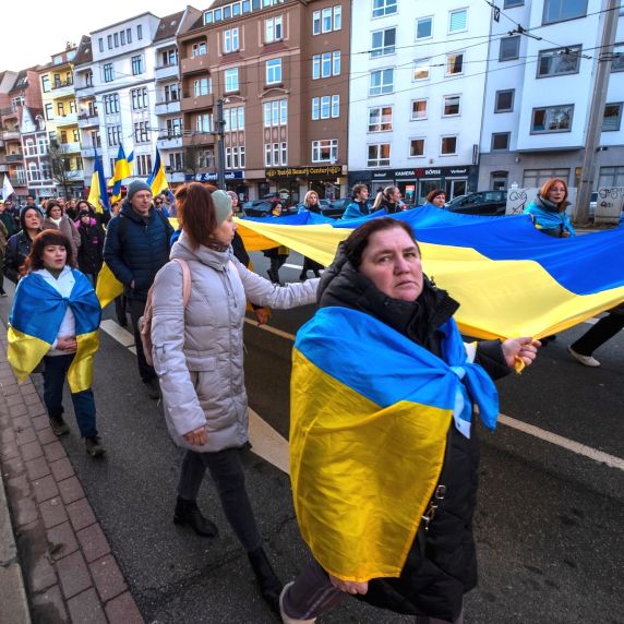 Menschen ziehen in einer Demonstration durch eine Stadtstraße und tragen eine große blau-gelbe Flagge.