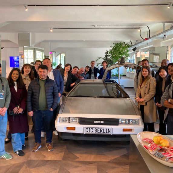 Eine Gruppe von Menschen posiert in einem hellen Innenraum um die Front eines DeLorean-Autos, dessen Kennzeichen „CIC BERLIN“ zeigt.