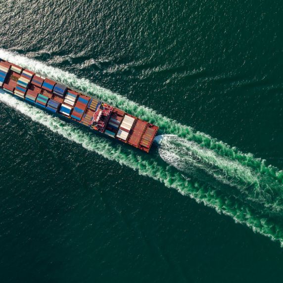 Luftaufnahme eines Containerschiffs, das über das Meer fährt und eine breite Bugwelle hinterlässt.