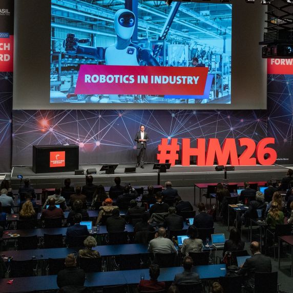Ein Mann steht auf einer Bühne. Hinter ihm an einer Leinwand ist groß der Schriftzug „Robotics in Industry“ zu lesen.