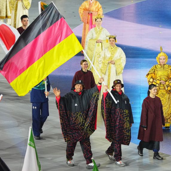 Die Rodler Tobias Wendl und Tobias Arlt tragen bei der Parade der Nationen eine große Deutschlandflagge und winken.