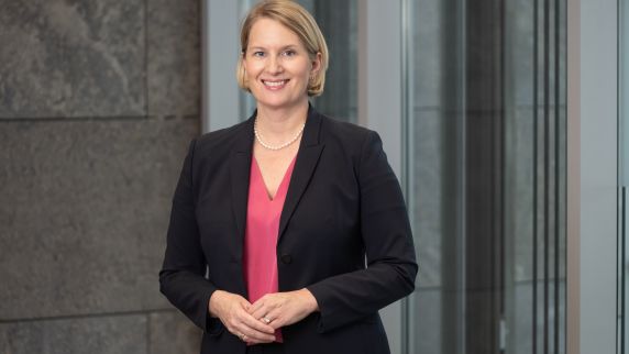 Professionelles Porträtfoto einer blonden Frau mit kinnlangem Haar, die einen schwarzen Blazer über einem roten Oberteil trägt und mit verschränkten Armen vor einem unscharfen grünen Hintergrund lächelt. 
