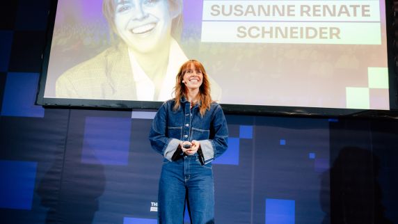 Eine lächelnde Frau mit Headset-Mikrofon steht auf einer Bühne und hält eine Fernbedienung, während hinter ihr auf einer großen Leinwand der Name „SUSANNE RENATE SCHNEIDER“ angezeigt wird.