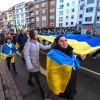 Menschen ziehen in einer Demonstration durch eine Stadtstraße und tragen eine große blau-gelbe Flagge.