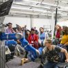 Eine Gruppe von Teilnehmenden mit blauen Namensbändern beobachtet in einem Robotiklabor der TU Darmstadt eine Demonstration, bei der ein blau-weißer kollaborativer Roboterarm einen Holzklotz greift, während ein junger Mann mit Brille und Laptop die Steuerung übernimmt. 