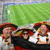 Vier Fußballfans in schwarz-rot-goldenen Hüten und mit Deutschland-Fanartikeln jubeln auf der Tribüne eines großen Stadions.