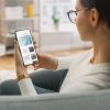 Young Woman at Home Uses Smartphone for Scrolling and Reading News about Technological Breakthroughs. She's Sitting On a Couch in Her Cozy Living Room. Over the Shoulder Shot