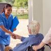 Senior Couple With Man In Wheelchair Greeting Female Nurse Or Care Worker Making Home Visit At Door