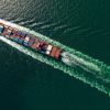 Luftaufnahme eines Containerschiffs, das über das Meer fährt und eine breite Bugwelle hinterlässt.