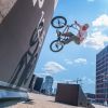 BMX-Fahrer springt mit dem Rad an einer Wandrampe hoch; im Hintergrund moderner Bürobau unter blauem Himmel.